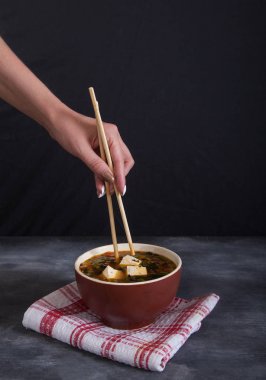 Siyah bir arka plan üzerinde kırmızı bir peçete üzerinde geleneksel Japon çorbası. Bir dişi el yemek çubukları ile tofu peyniri alır. Dikey
