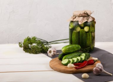 Cam kavanozda mayalanmış salatalıklar, ahşap bir tahtanın üzerinde taze salatalıklar, sıcak kırmızı biber, sarımsak, dereotu, beyaz ahşap arka planda gri peçete..