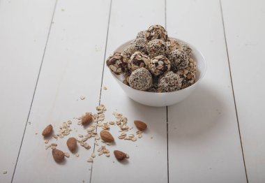 Enerji dolu hurma topları ve susam tohumlu bademler beyaz bir porselen kabın içinde uzanır, yanında beyaz bir masanın üzerine yayılmış bademler vardır. Faydalı fast food.