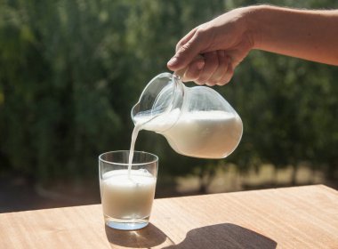 Erkek el, cam sürahisinden yeşil ağaçların arka planındaki bir cama kefir döker. Fermente edilmiş ürün.