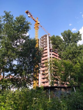 Uzun bir vinç, yazın mavi gökyüzüne karşı yeşil ağaçların arasına bir ev inşa ediyor. Mimarlık. Ekoloji. dikey.