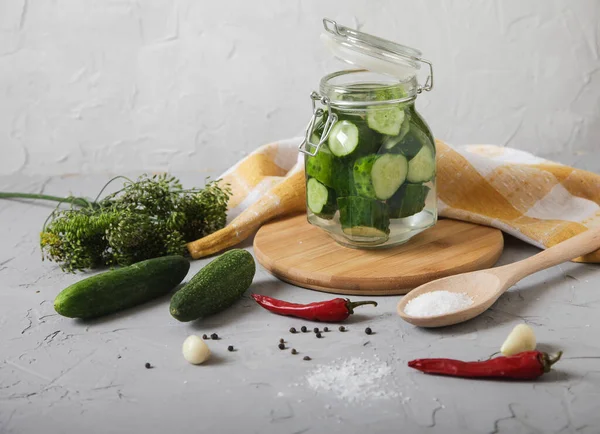 Küçük cam bir kavanozda parçalanmış salatalık, kapak, biber, ahşap kaşıkta tuz beton bir arka planda duruyor..