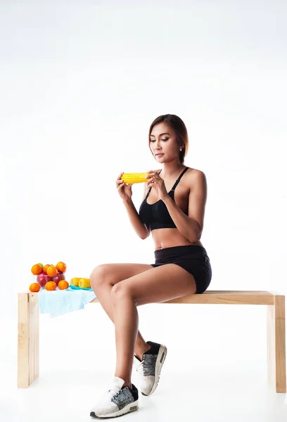 The lady in exercise suit is sitting on wooden chair,with fruit bowl,red apple and orange in bowl, she is hold yellow corn in her hands,prepare for eat,fresh fruit ,good for health and life