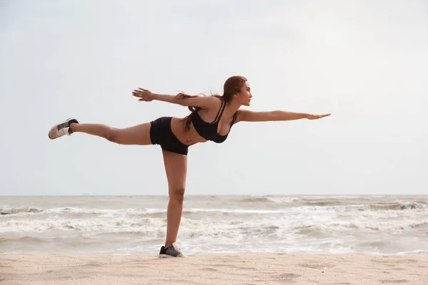lady with sportwear is stand with left leg side and raise right leg up in the air,raise left hand up in the air and bend like arrow ,posing yoga pattern on the beach