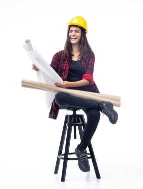 Genç kadın mühendis kafasına sarı kaskı taktı, siyah tahta sandalyeye oturdu, elindeki planlara baktı, gülümseyerek ve mutlu bir şekilde.,