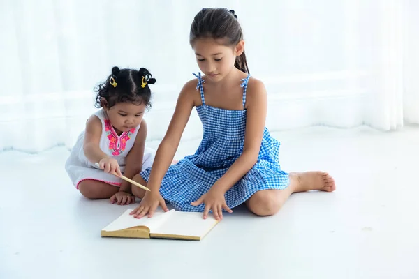 The elder sister opening book,younger sister sitting beside her and using pencil write on book,doing activity together,at home — Stock Image Elder Sister Opening Book Younger Sister Sitting Her Using Pencil Stock Photo