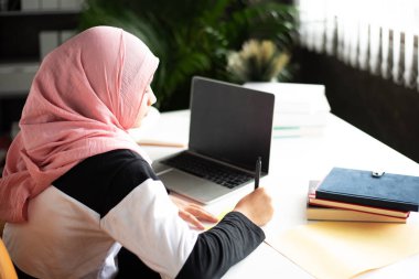 Müslüman kadın kağıda yazıyor, laptoptan veri kaydediyor, evde çalışıyor, dizüstü bilgisayarını ve kitaplarını ahşap masaya koyuyor.,