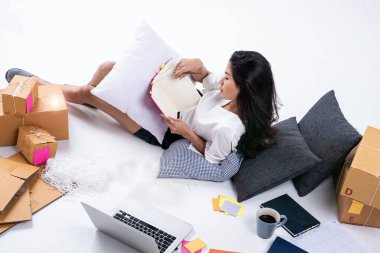 Posta kutusunun yanına uzanmış güzel bir bayan, dizüstü bilgisayar ve kitap kullanarak çevrimiçi satış, gönderme için paketleme, evde çalışma, e-ticaret, mercek efekti gibi bilgileri kaydediyor.
