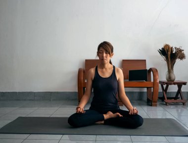 Egzersiz kıyafeti giyen, yoga minderinde oturan, gözlerini kapayan, meditasyon yapan, evde yoga yapmaya hazırlanan, dinlenme zamanı