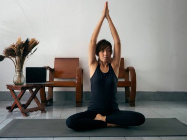 Egzersiz kıyafeti giyen bir kadın, paspasın üzerine oturur, el ele tutuşur ve başını kaldırır, gözlerini kapatır, yogadan önce meditasyon yapar.