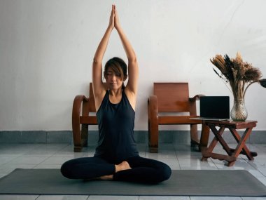 Egzersiz kıyafeti giyen bir kadın, paspasın üzerine oturur, el ele tutuşur ve başını kaldırır, gözlerini kapatır, yogadan önce meditasyon yapar.