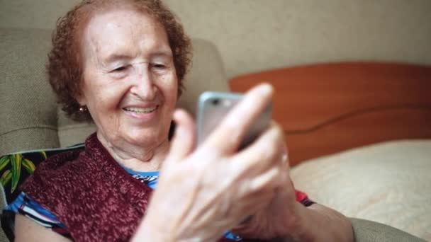 An old woman writes a message and looks at the photos on her new smartphone. Grandma with deep wrinkles. Indoors. Happy senior woman uses smartphone sitting on sofa at home. Happy old age. Portrait. – stockvideo