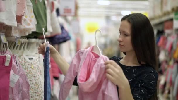 Piękna dziewczyna wybierając ubrania dla dziecka stojącego w pobliżu półki odzieży dla dzieci w supermarkecie, zakupy, magazyn centrum handlowego. Kobieta kupuje różowe ubrania na świeżym powietrzu dla dziecka na sprzedaż w sklepie ubrania — Wideo stockowe