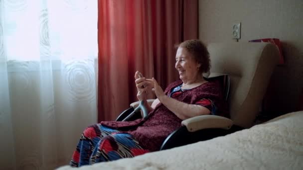 Gammal kvinna skriver ett meddelande och tittar på bilderna på sin smartphone. Mormor med djupa rynkor. Inomhus. Glad äldre kvinna använder smartphone sitter på soffan hemma. Lycklig ålderdom. Porträtt. — Stockvideo