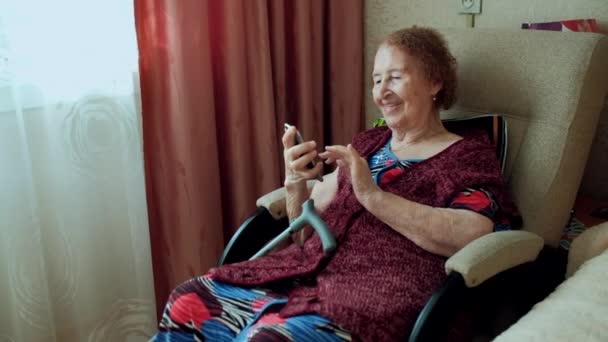 En gammal kvinna skriver ett meddelande och tittar på bilderna på sin nya smartphone. Mormor med djupa rynkor. Inomhus. Glad äldre kvinna använder smartphone sitter på soffan hemma. Porträtt. — Stockvideo