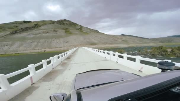 Auto podróż: Suv jeździ po wysokiej górskiej drodze na białym moście nad rzeką. Pov - Punkt widzenia samochodu poruszającego się po drodze w górach do przodu wsi. Podróże off road. Koncepcja podróży samochodem. — Wideo stockowe