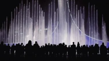 İnsanlar arka plandaki parlak renk çeşmesinde siluetler oluşturuyor. Bir grup insan şehir gece görüşünü, ışığı ve müzik fıskiyesini seyrediyor. Turizm şehirleri
