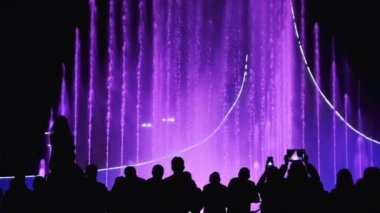 Bir grup insan şehir gece görüşünü, ışığı ve müzik fıskiyesini seyrediyor. İnsanlar arka plandaki parlak renk çeşmesinde siluetler oluşturuyor. Turizm şehirleri 