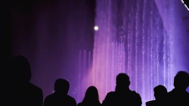 İnsan siluetleri. Işık festivali sırasında renkli çeşmeler. Bir grup insan şehrin gece manzarasının, ışığın ve müzik çeşmesinin tadını çıkarıyor.. 