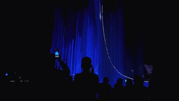 Gruppenmenschen genießen den nächtlichen Blick auf die Stadt, das Licht, die Springbrunnenshow. Ansicht von Menschen Silhouetten mit dem Smartphone Fotos von tanzenden Springbrunnen zeigen. — Stockvideo
