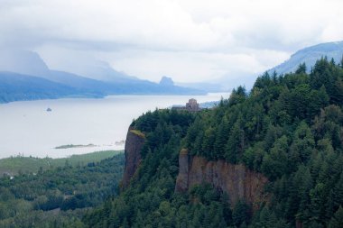 Uzak manzara ev - Columbia river gorge. Kayalık tepenin üstüne bina dönüm noktası. Bulutlar ve ufuktaki dağlar. Yeşil orman her yerde.