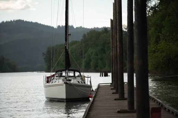 Docked sailboat/yacht at the wooden pier/dock with wooden poles on the right. Boat on Willamette river with mountains and sky in the background