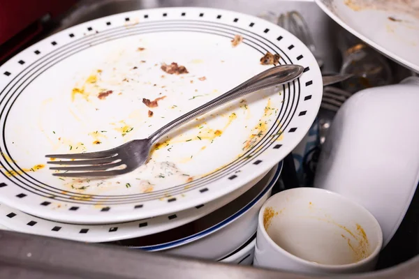 Pile of dirty dishes in the sink. Plate with a fork on top with leftovers of eggs and bacon.