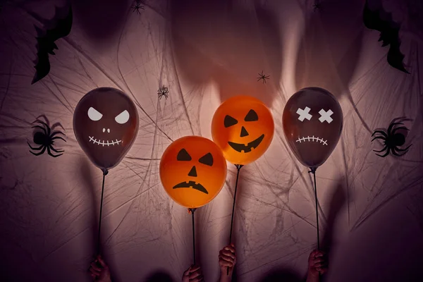 Halloween balloons background with horror faces held by 7 year old children\'s hands. Dark background with shadows, spiders, spiderwebs and bats.