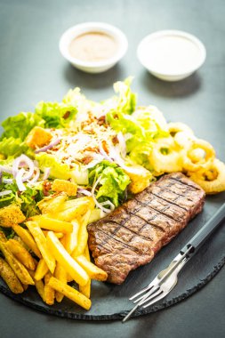 Sığır eti biftek ile kızarmış patates soğan halkası sos ve taze sebze siyah Arduvaz ile ızgara