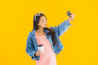 Kahve fincanı ve sarı izole edilmiş arka planda akıllı cep telefonlu güzel Asyalı kadın portresi.