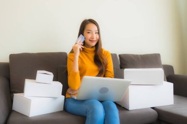 Asyalı genç bir kadının portresi, oturma odasındaki koltukta paket kutusuyla çalışmak için dizüstü bilgisayar kullanıyor.