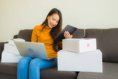 Asyalı genç bir kadının portresi, oturma odasındaki koltukta paket kutusuyla çalışmak için dizüstü bilgisayar kullanıyor.