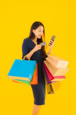 Portre güzel genç Asyalı kadın alışveriş çantası kredi kartı ve akıllı cep telefonu sarısı izole bir arka plan.