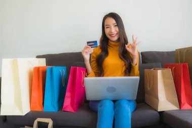 Genç Asyalı kadının oturma odasında online alışveriş yapmak için akıllı cep telefonu ve kredi kartı olan dizüstü bilgisayarı kullandığı portre.