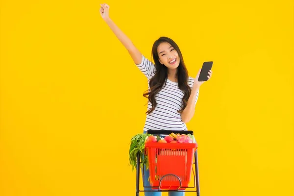 ポートレート美しい若いアジアの女性スーパーマーケットから食料品を買い物し 黄色の隔離された背景にスマートフォンでカート — ストック写真
