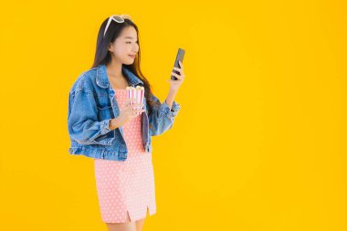 Güzel bir Asyalı kadın portresi telefon patlamış mısırı ve sarı izole edilmiş bir arka planda film izlemekten memnun.