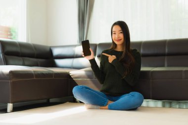 Güzel bir Asyalı kadın portresi oturma odasının iç tarafında kanepeli bir kahve fincanı ve akıllı cep telefonu kullanıyor.