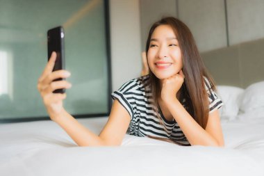 Güzel bir Asyalı kadın yatak odasının içinde akıllı cep telefonu kullanıyor.