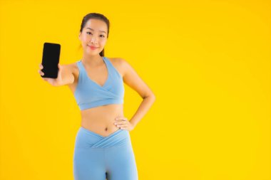 Güzel bir Asyalı kadın portresi sarı izole edilmiş arka planda akıllı cep telefonuyla spor giyer.