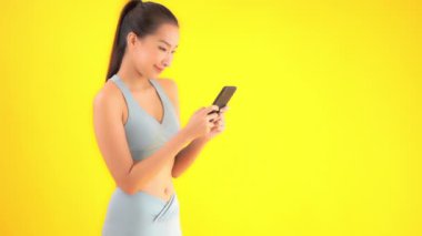 Sarıya izole edilmiş akıllı telefonlu, spor tişörtlü güzel Asyalı bir kadının görüntüleri.