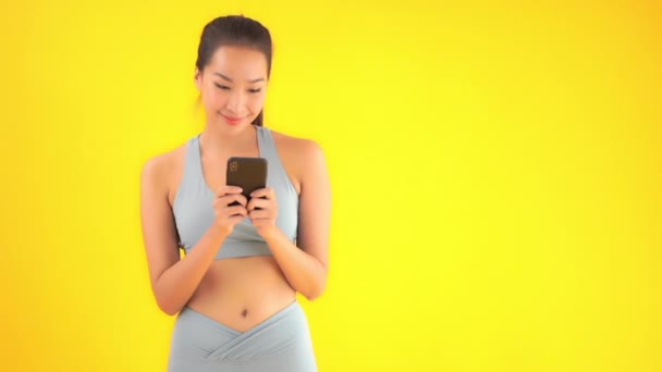 Zdjęcia Pięknej Młodej Azjatyckiej Kobiety Bluzce Sportowej Smartfonem Odizolowanym Żółto — Wideo stockowe