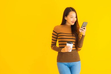 Güzel, genç Asyalı bir kadın sarı izole edilmiş arka planda kahve fincanı ile akıllı cep telefonu veya cep telefonu kullanır.