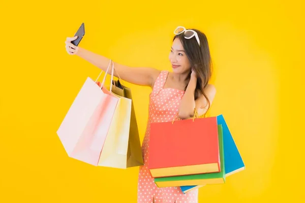 亚洲年轻貌美的女子 头戴五颜六色的购物袋 手持智能手机 背景为黄色 背景为孤立的信用卡 — 图库照片