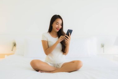 Güzel bir Asyalı kadın portresi. Yatak odasında beyaz yastık battaniyesi ve yatağında akıllı cep telefonu kullanıyor.