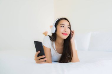Asyalı güzel bir kadının portresi mutlu bir gülümseme akıllı cep telefonu ve kulaklıkla yatak odasının içinde müzik dinlemek için.
