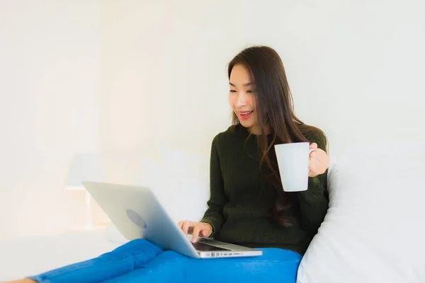 在卧室内带着手提电脑和咖啡杯在床上工作的漂亮年轻亚洲女人的画像 — 图库照片