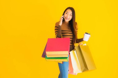 Güzel, genç Asyalı bir kadın sarı izole arka planda kahve fincanı ve renkli alışveriş çantasıyla akıllı cep telefonu veya cep telefonu kullanır.