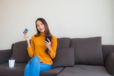 Genç Asyalı kadının oturma odasında online alışveriş yapmak için akıllı cep telefonu ve kredi kartı olan dizüstü bilgisayarı kullandığı portre.