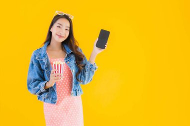 Güzel bir Asyalı kadın portresi telefon patlamış mısırı ve sarı izole edilmiş bir arka planda film izlemekten memnun.