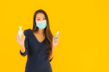 Maske, alkol jeli ve sarı izole edilmiş arka planda spreyli güzel Asyalı kadın portresi.